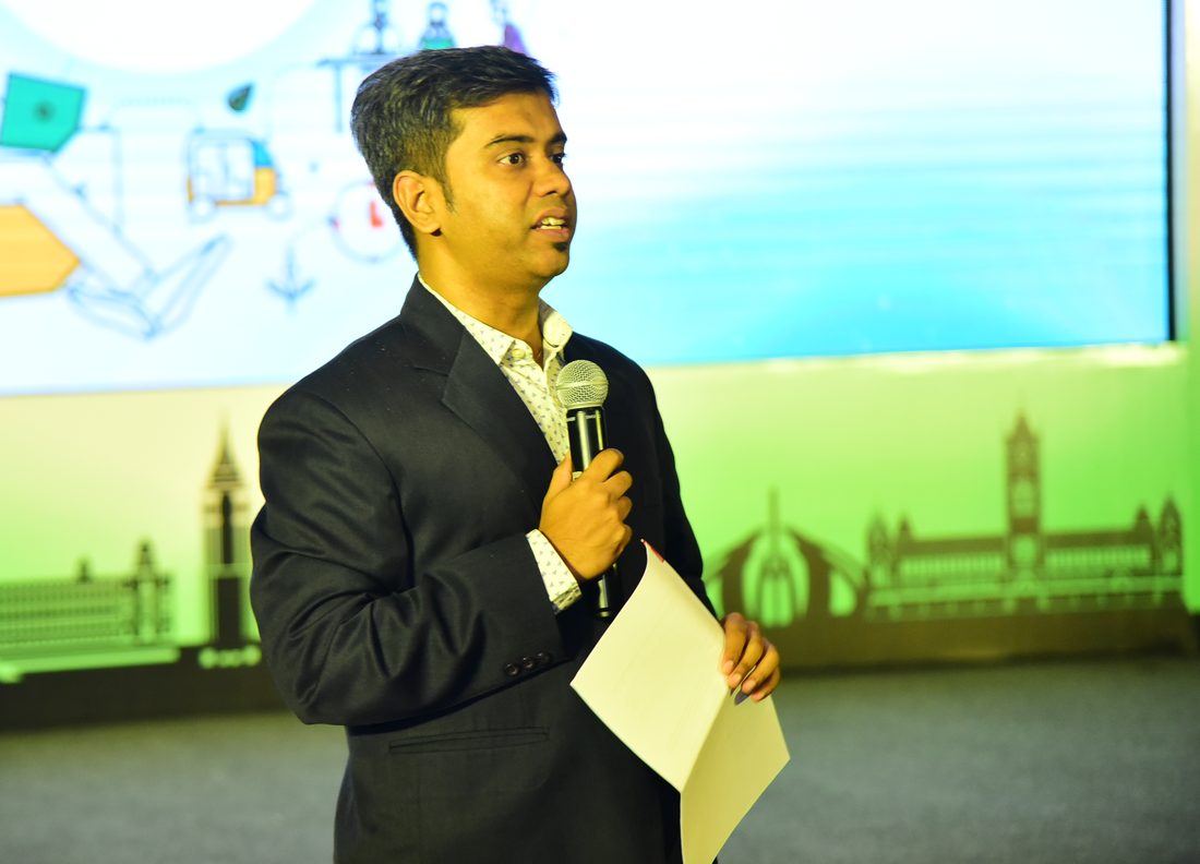 Parijat Sarkar on stage in a dark suit, microphone in one hand and his notes in the other